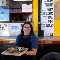 young hispanic woman standing in front of food truck with a tray of tacos